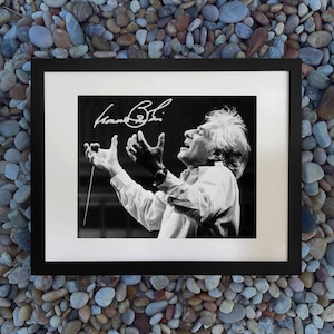 May include: Framed black and white photograph of a conductor with arms raised, wearing a white shirt and wristwatch. The image includes a signature in the upper left corner. The photograph is set against a white mat and a black frame.