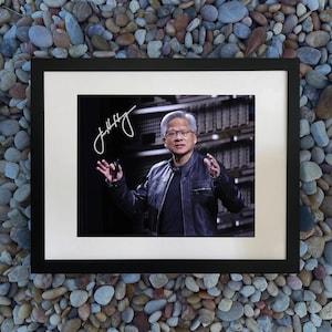 May include: Framed print featuring a man in a black leather jacket, glasses, and a black shirt. The print has a white border and a black frame. The man is gesturing with his hands. The print is signed in white ink.