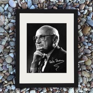 May include: Black and white framed portrait of an older man wearing glasses, a suit, and tie. The man is looking to the left, with his hand on his chin. The frame is black, and the background is a mix of pebbles.