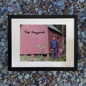 May include: Framed photograph featuring a man standing in front of a small wooden cabin. The cabin is painted a reddish-brown color with a green roof. The photograph is signed "Ted Kaczynski" in black ink.