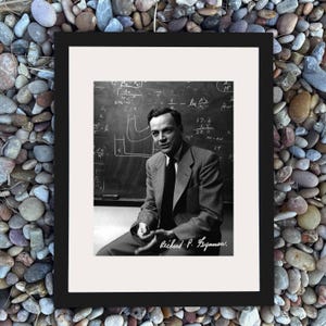 May include: Black framed print showing a black and white photograph of Richard Feynman. He is seated in front of a chalkboard with equations, wearing a suit and tie. The print is set against a background of small, colourful stones.