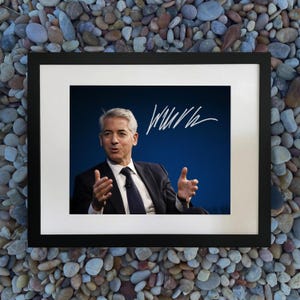 Puede incluir: Una fotografía enmarcada que presenta a un hombre con traje oscuro y corbata, gesticulando con las manos. El fondo es azul oscuro, con escritura blanca en la esquina superior derecha. El marco es negro y el fondo es un lecho de pequeñas piedras de colores.