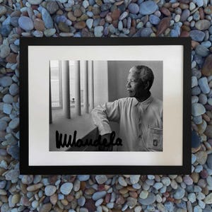 May include: Black and white framed print featuring a portrait of Nelson Mandela. The image shows Mandela in a light-colored shirt, leaning against a railing, with his signature at the bottom. The background includes pillars and a view of the outside.