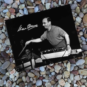 May include: Black and white photograph of a man conducting an orchestra. The man is wearing glasses and a short-sleeved shirt. The photograph is signed "Mar Stevens". The image is resting on a bed of colorful pebbles.