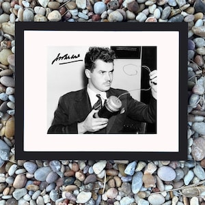 May include: Black and white framed photograph of a man in a suit holding a cylindrical object with wires. The photograph has a signature in the upper left corner. The frame is black, and the background is a collection of small, smooth stones.
