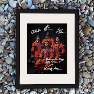 May include: Framed print featuring a group of five astronauts in orange space suits. The print includes signatures and the text "Back to the Moon with Artemis". The astronauts are posed against a dark background.