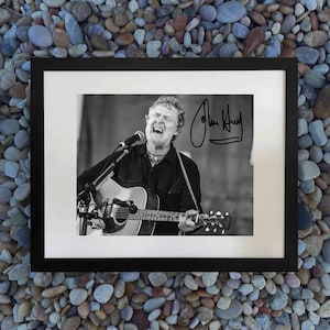 May include: A black and white framed photograph of a musician with a beard singing into a microphone while playing an acoustic guitar. The image features a signature in the upper right corner and is enclosed in a simple black frame with a white mat.