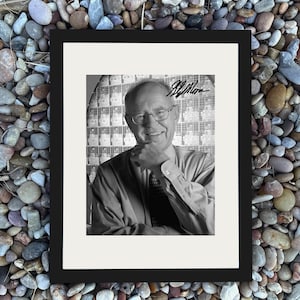 May include: Black and white framed portrait of a smiling man wearing glasses and a collared shirt. The background features a grid-like pattern. The frame is black, and the portrait is signed in the upper right corner.