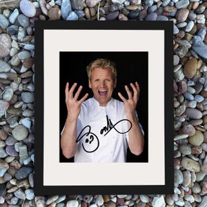 May include: Framed photograph featuring a man in a white chef's coat, with his hands raised and mouth open in an expression of excitement. The image is signed with a black marker. The frame is black, and the background is a collection of pebbles.
