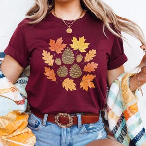 Puede incluir: Camiseta burdeos con un diseño circular de hojas de otoño, piñas y bellotas. Las hojas son en tonos naranja, amarillo y marrón. La camiseta tiene las mangas dobladas y se combina con pantalones cortos vaqueros azules y un cinturón marrón.