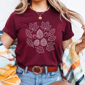 Puede incluir: Camiseta burdeos con un diseño blanco de hojas de otoño, piñas y bellotas. La camiseta de manga corta tiene las mangas enrolladas y se combina con pantalones cortos vaqueros azules y un cinturón marrón con hebilla decorativa. Se ve un collar dorado.