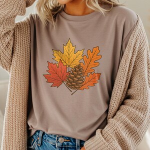 Puede incluir: Una camiseta color topo con un diseño de temática otoñal. El diseño incluye un grupo de hojas de otoño en tonos rojos, naranjas y amarillos, junto con una piña marrón. Las hojas y la piña están delineadas en negro.