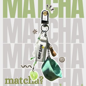 Puede incluir: Un llavero de metal plateado que presenta un dije de cartón blanco con la etiqueta OATSIDE, un cuenco de cerámica verde claro, un pequeño dije de batidor marrón y una cuenta translúcida en forma de corazón verde, unido a una cadena.