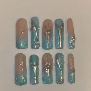 May include: A set of ten artificial nails. Each nail features a light pink base with a blue tip. Some nails have silver metallic embellishments, including spider-like designs and abstract shapes.