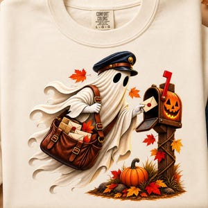 Peut inclure: Un sweat-shirt à col rond couleur crème avec une illustration représentant un fantôme portant une casquette de facteur et une sacoche en cuir marron. Le fantôme dépose une lettre dans une boîte aux lettres en bois en forme de citrouille d'Halloween.