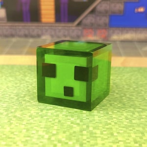 Peut inclure: Figurine cubique verte translucide avec des yeux et une bouche carrés noirs. La figurine présente un design pixélisé, rappelant un personnage de jeu vidéo. L'arrière-plan est flou, avec une surface verte pixélisée.