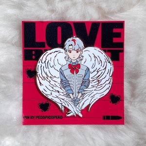 May include: A red enamel pin featuring a character with white wings and the text "LOVE". The character has white hair with red accents, a blue uniform, and a red bow tie. The pin is on a red background with black hearts and a bullet.