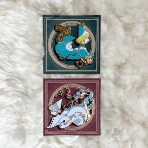 May include: Two enamel pins on cards. The top pin features a character in teal and gold, with the name "Coco" above. The bottom pin shows a character in white and blue, with the name "Oifrey" above. Both pins have gold accents and are set against a white, fluffy background.