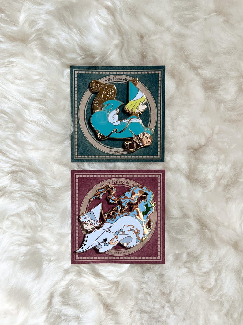 May include: Two enamel pins on cards. The top pin features a character in teal and gold, with the name "Coco" above. The bottom pin shows a character in white and blue, with the name "Oifrey" above. Both pins have gold accents and are set against a white, fluffy background.