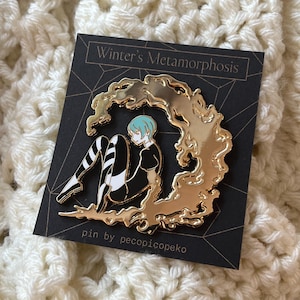 May include: A gold enamel pin depicting a seated figure with blue hair and black and white striped legs, set against a gold cloud design. The pin is on a black card with the text "Winter's Metamorphosis" and "pin by pecopicopeko".