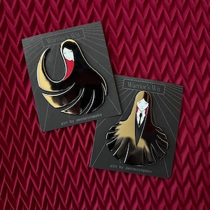 May include: Two enamel pins featuring a stylized female figure with long black hair and gold accents. The pins are on black cards with the text "Warrior's Wit" and "pin by pecopicopeko". The background is a textured burgundy fabric.