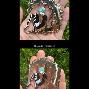 May include: Gold-colored metal pin featuring a seated anime-style figure with blue hair and black and white striped legs. The figure is surrounded by a stylized, flowing gold design. The pin is held in a hand.