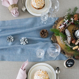 Può includere: Una tavola apparecchiata per le feste con un runner in lino blu, due coperti con tovaglioli rosa e una fetta di legno con una ghirlanda natalizia. Ogni coperto comprende un piatto bianco con un dolce ricoperto di mandorle e zucchero a velo, un cucchiaio d'argento e un bicchiere. La tavola è decorata con pigne e un piccolo cartello di legno con il nome "Kate" scritto su di esso.