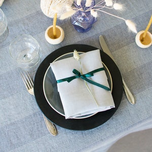 Può includere: Una mise en place con un piatto nero e grigio, un tovagliolo di lino bianco legato con un nastro verde, un bicchiere d'acqua, una forchetta e un coltello. La tavola è coperta da una tovaglia a quadri grigi e bianchi.