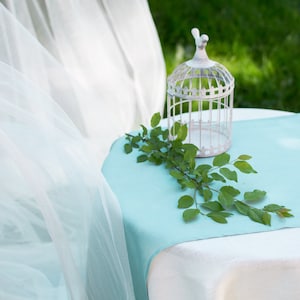 May include: A white birdcage with a bird on top sits on a light blue table runner on a white tablecloth. Green leaves are arranged on the table runner.