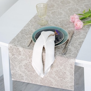 以下が含まれることがあります： ベージュのダマスク柄テーブルランナー、ターコイズブルーのプレート、白い折り畳まれたナプキン、透明なグラスが特徴のテーブルセッティング。ピンクのチューリップと銀色のナイフがエレガントなアレンジを完成させています。