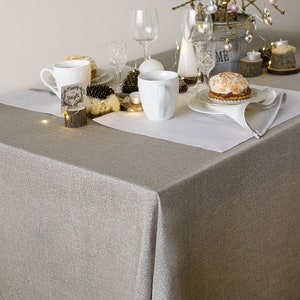 May include: A white tablecloth with a silver shimmer covers a table set for a festive meal. A white mug with a handle sits on a white saucer. A small wooden sign with the text "Merry & Bright" sits on a small wooden log. A white plate with a pastry sits on a white napkin. A small, twig Christmas tree with pink and silver baubles sits in a metal pot.