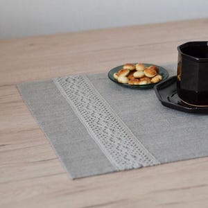 Puede incluir: Un mantel individual gris con un diseño de encaje blanco, una taza y un platillo negros y un plato pequeño con galletas. La taza y el platillo tienen un diseño geométrico. El mantel individual está sobre una mesa de madera.