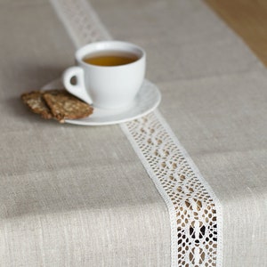 May include: A white linen table runner with a white lace trim. A white cup of tea and a plate of cookies are on the table runner.