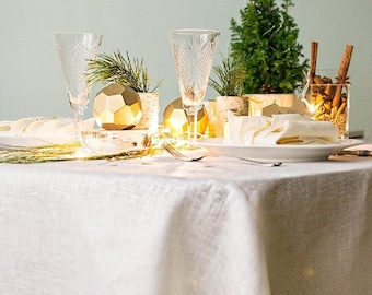 Könnte beinhalten: Eine weiße Leinen-Tischdecke mit einem goldenen geometrischen Ornament, zwei klaren Gläsern und einem kleinen Topf-Evergreen-Baum. Der Tisch ist für ein festliches Abendessen mit einer weißen Serviette und einem weißen Teller gedeckt.