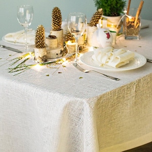 May include: A white linen tablecloth with a subtle texture, set for a holiday dinner. The table is decorated with pine sprigs, pinecones, and a string of fairy lights. A white plate with a white napkin is set on the table.