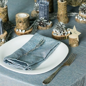 Può includere: Una mise en place blu con un piatto bianco, un tovagliolo blu, una forchetta d'argento e un segnaposto in legno con il nome "Tom" su di esso. Il tavolo è decorato con pigne argentate, portacandele in legno e rametti di fogliame argentato.