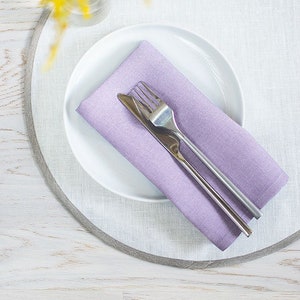 Peut inclure: Une assiette blanche avec une serviette en lin violette et une fourchette et un couteau en argent sur le dessus. L'assiette est sur un dessous de plat rond gris clair.