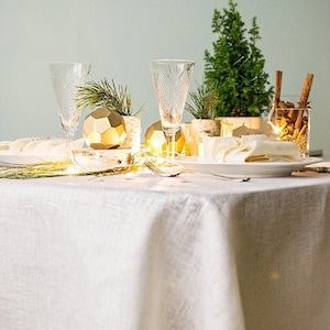 Può includere: Una tovaglia di lino bianco con un ornamento geometrico dorato, due bicchieri trasparenti e un piccolo albero sempreverde in vaso. La tavola è apparecchiata per una cena festiva con un tovagliolo bianco e un piatto bianco.