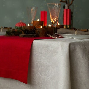 Può includere: Una tovaglia di lino bianca con un delicato motivo floreale, un runner rosso e una mise en place con un piatto bianco, posate e un bicchiere di vino. Il tavolo è decorato con candele e verde.