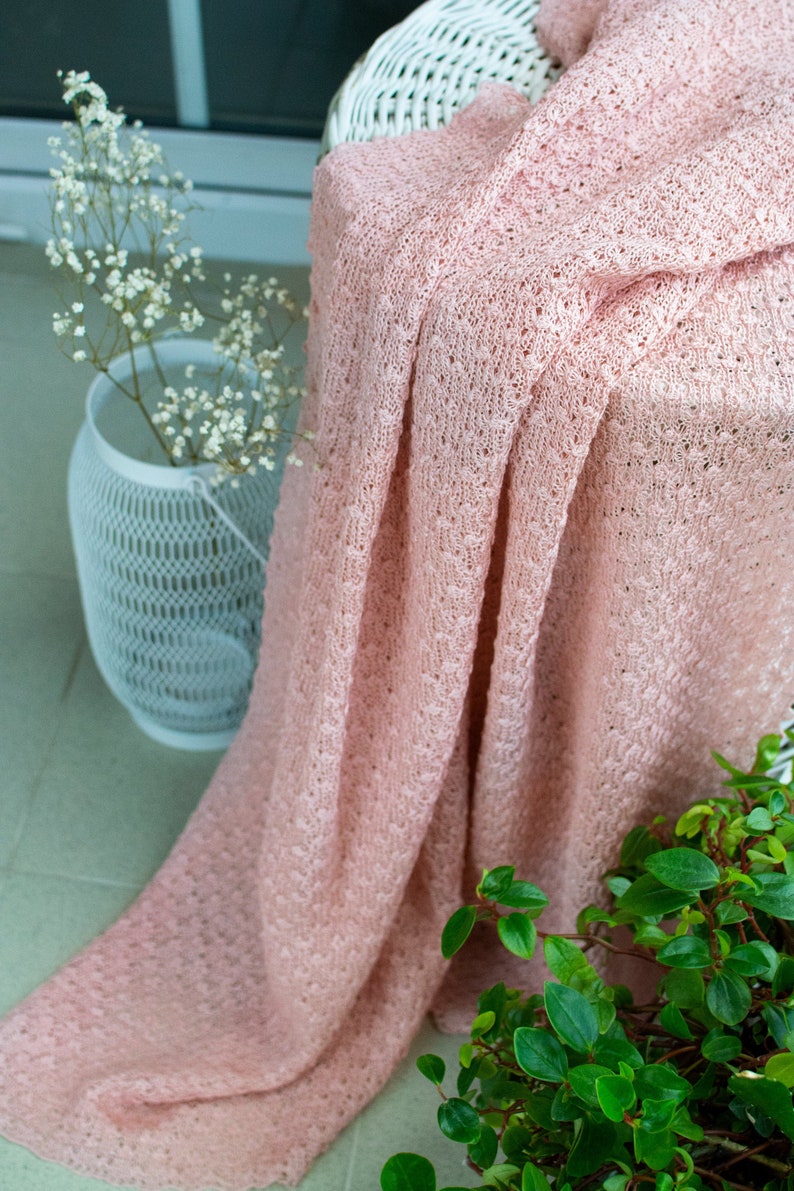 K&ouml;nnte beinhalten: Eine weiche, rosafarbene gestrickte Wolldecke, die &uuml;ber einen wei&szlig;en Korbstuhl gelegt ist. Die Decke hat ein zartes, offenes Webmuster und ist von gr&uuml;nen Pflanzen umgeben.