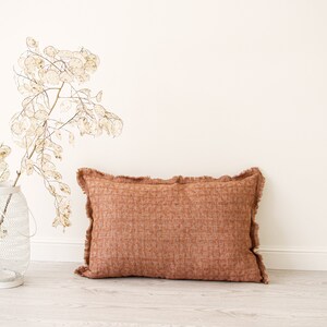 May include: A rectangular, burnt orange throw pillow with a fringed edge. The pillow is lying on a light wood floor.