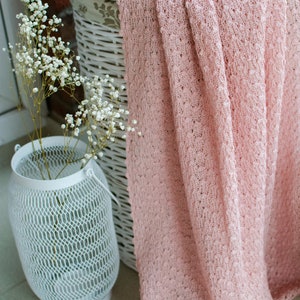 K&ouml;nnte beinhalten: Ein wei&szlig;er Korb aus Weide mit einem rosa gestrickten &Uuml;berwurf, der dar&uuml;ber gelegt ist. Der Korb steht auf einem gefliesten Boden neben einer wei&szlig;en Metall-Laterne mit einem Blumenarrangement.