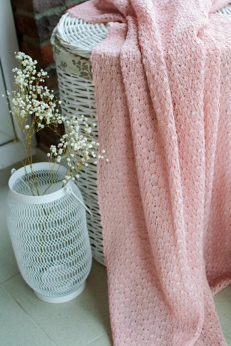 K&ouml;nnte beinhalten: Ein wei&szlig;er Korb aus Weide mit einem rosa gestrickten &Uuml;berwurf, der dar&uuml;ber gelegt ist. Der Korb steht auf einem gefliesten Boden neben einer wei&szlig;en Metall-Laterne mit einem Blumenarrangement.