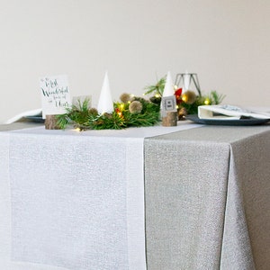 May include: A white linen table runner with a silver glitter tablecloth underneath. The table is set with a centerpiece of pine branches, white paper cones, and a string of fairy lights. A small wooden sign with the text "Most Wonderful Time of the Year" is placed on the table.