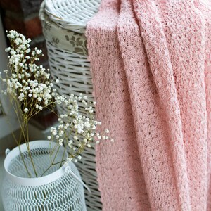K&ouml;nnte beinhalten: Ein wei&szlig;er Weidenkorb mit einer rosa geh&auml;kelten Decke dar&uuml;ber. Ein Strau&szlig; wei&szlig;er Babyschnabelblumen steht in einer wei&szlig;en Metallvase.