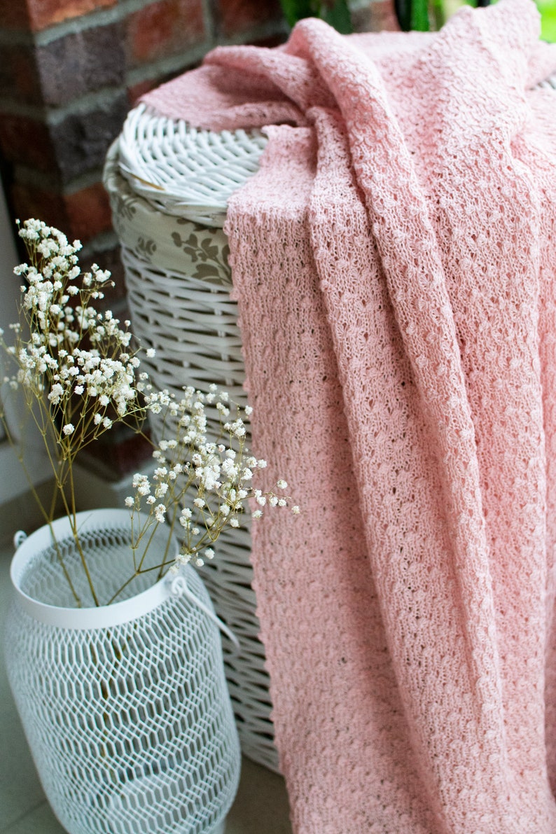 K&ouml;nnte beinhalten: Ein wei&szlig;er Weidenkorb mit einer rosa geh&auml;kelten Decke dar&uuml;ber. Ein Strau&szlig; wei&szlig;er Babyschnabelblumen steht in einer wei&szlig;en Metallvase.