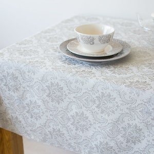 May include: White tablecloth with a silver damask pattern. A white teacup and saucer are on top of the tablecloth.