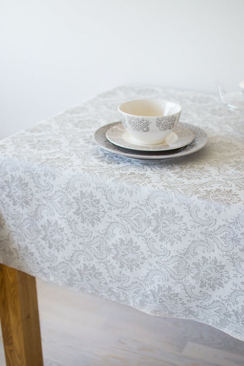 May include: White tablecloth with a silver damask pattern. A white teacup and saucer are on top of the tablecloth.