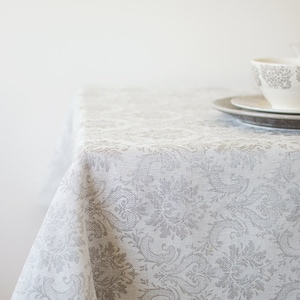 May include: A white tablecloth with a gray floral pattern. The tablecloth is draped over a table and has a slight wrinkle in the center.