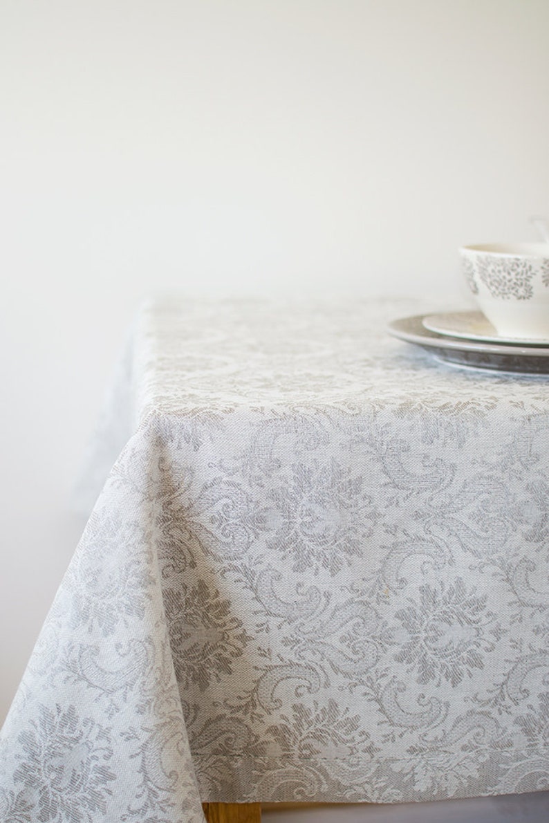 May include: A white tablecloth with a gray floral pattern. The tablecloth is draped over a table and has a slight wrinkle in the center.